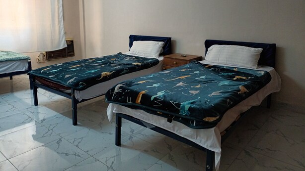 Una habitación con dos camas individuales, cada una con una colcha oscura estampada y almohadas blancas. Hay una mesita de noche de madera entre las camas y una ventana con cortina en el lado izquierdo.