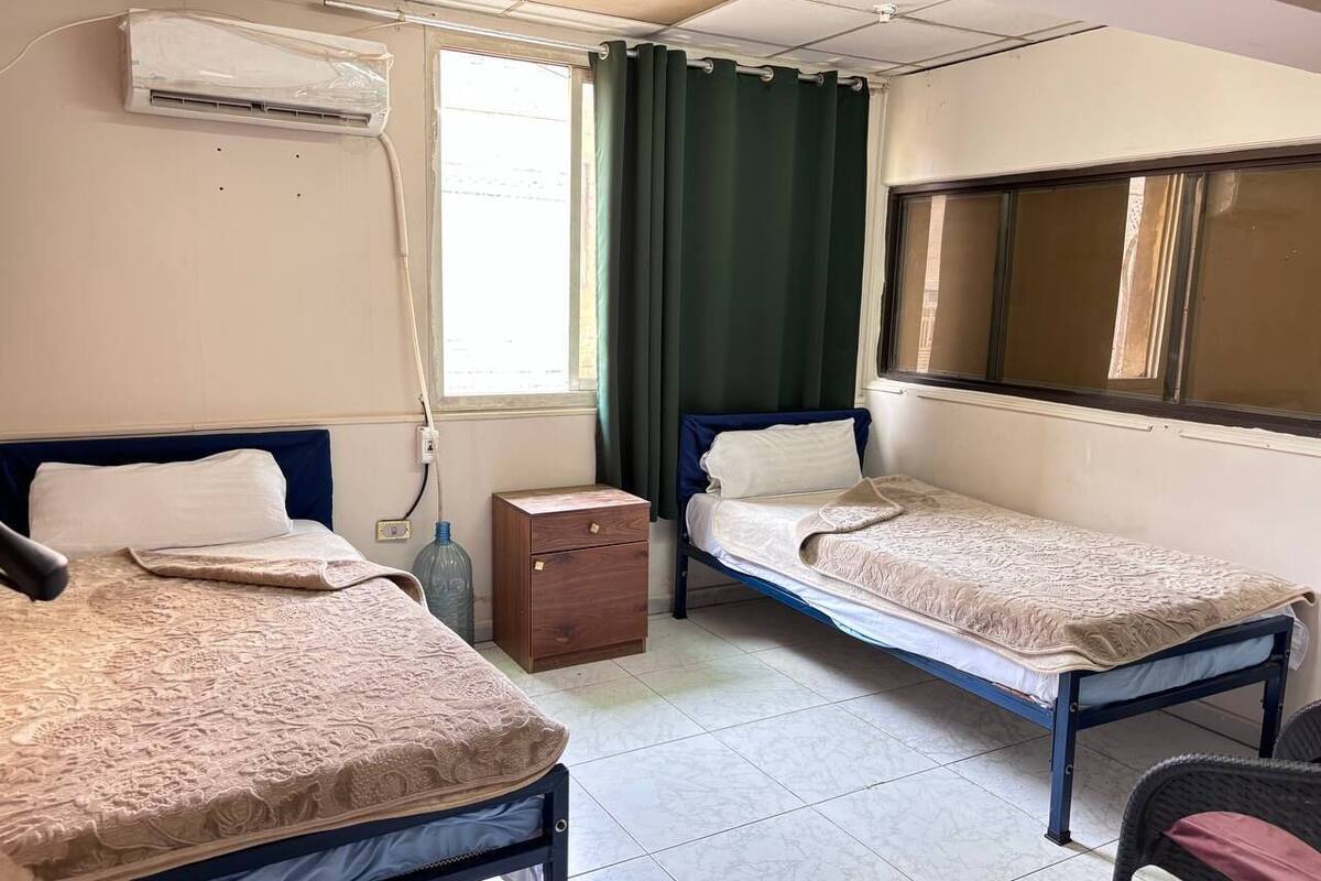 A small, clean room with two single beds, a wooden nightstand, a wall-mounted air conditioner, and a window with curtains.