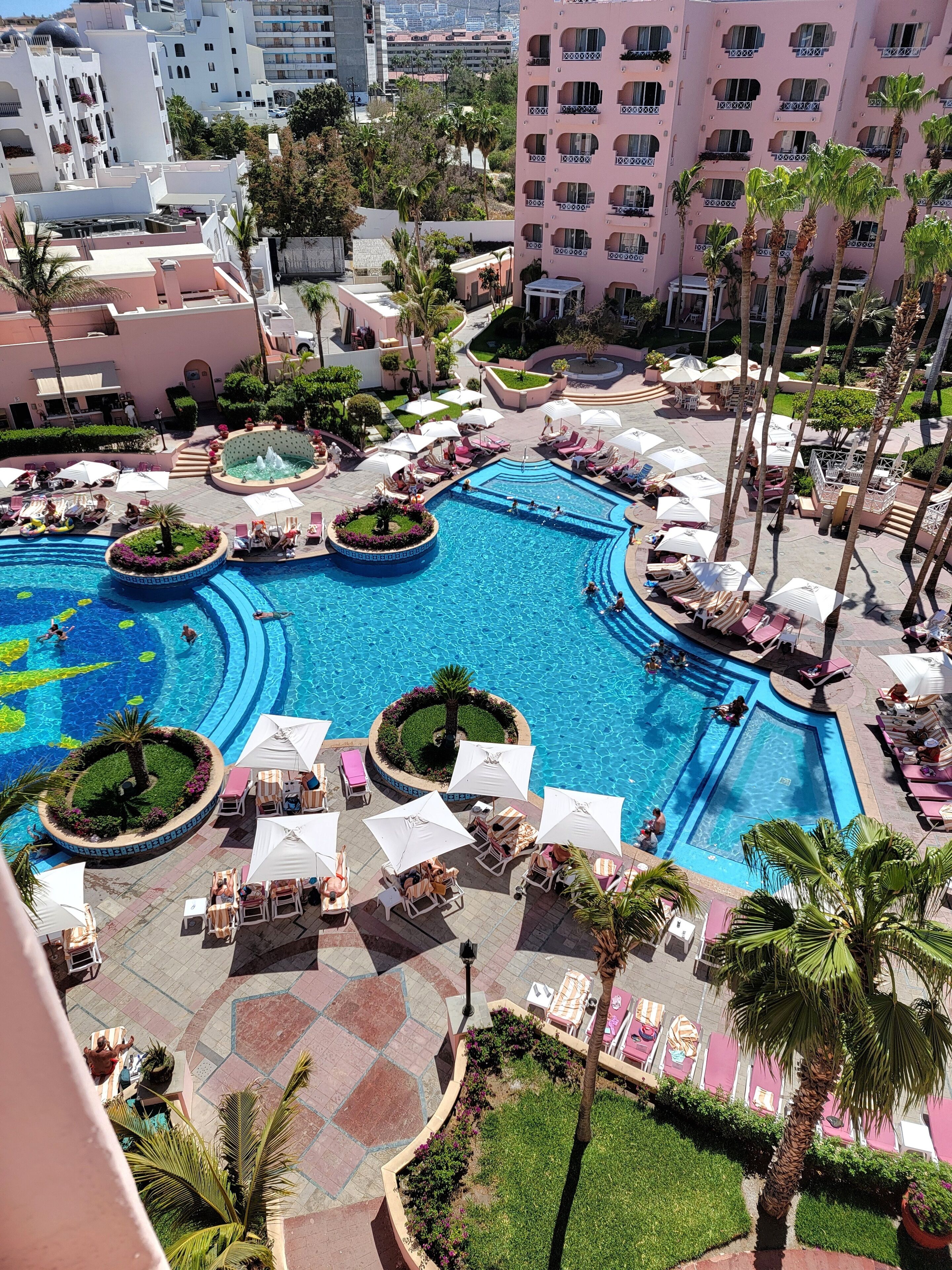 Zona de piscina en un complejo turístico, con varias piscinas, tumbonas y palmeras.