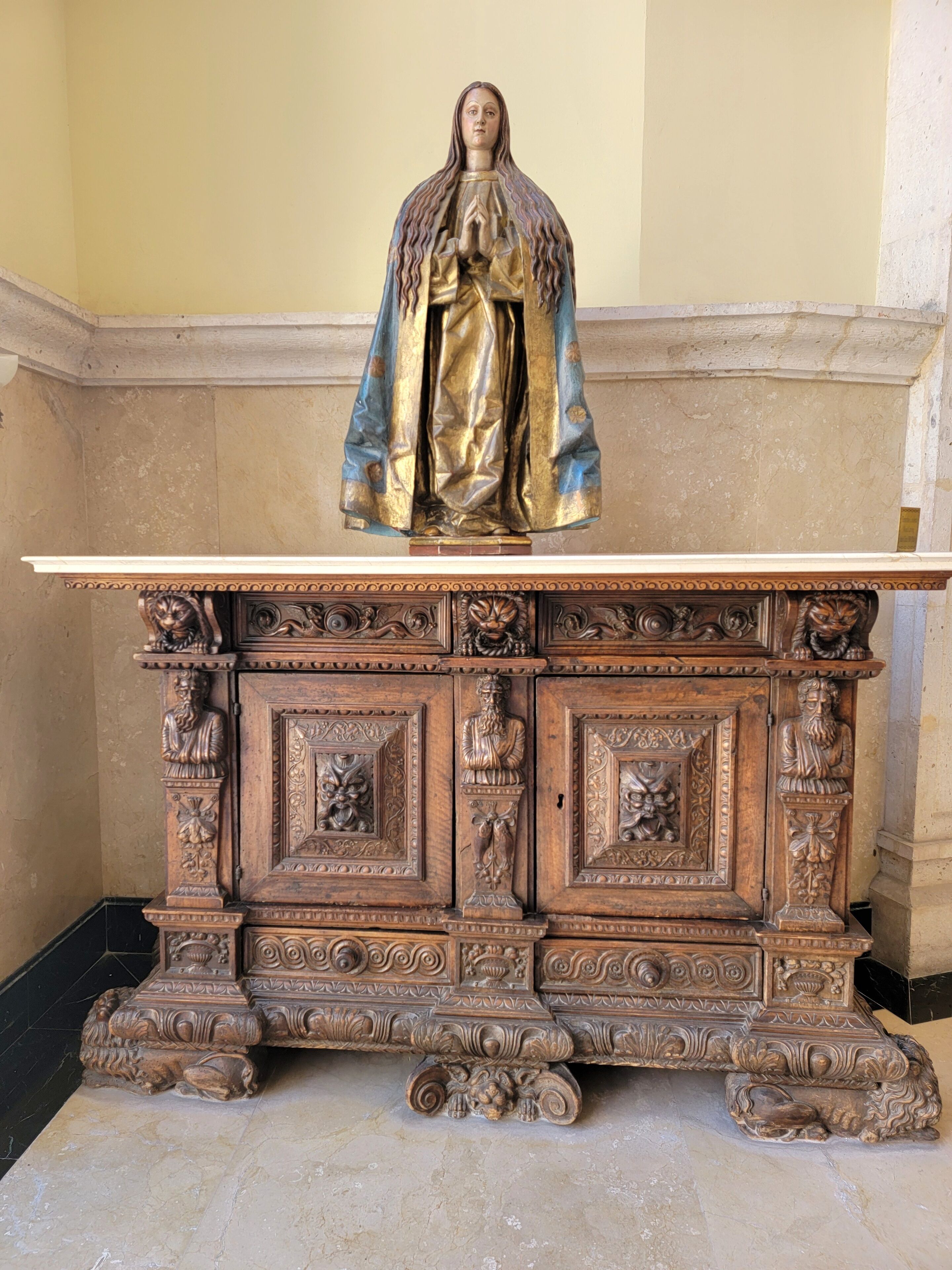 Una estatua de una figura vestida sobre un altar de madera con tallas intrincadas y una base de mármol.