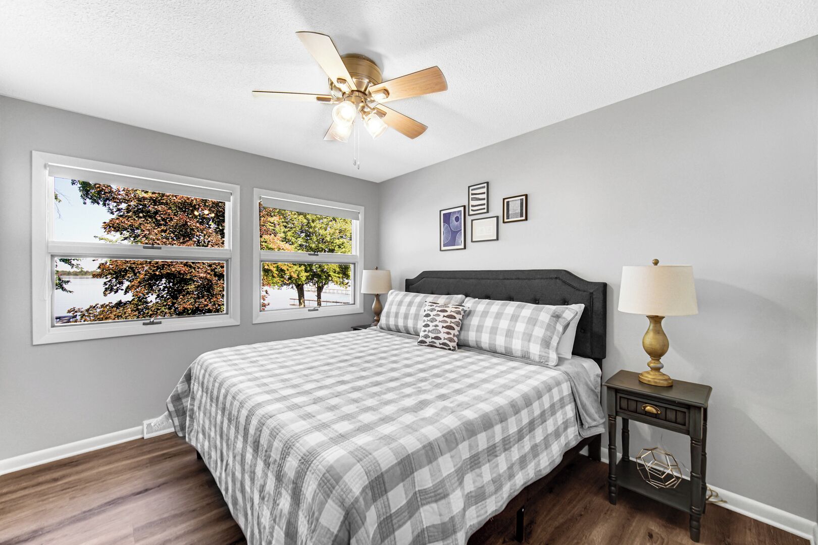 A bedroom with a bed, two bedside tables, a ceiling fan, and three windows with autumn foliage views.