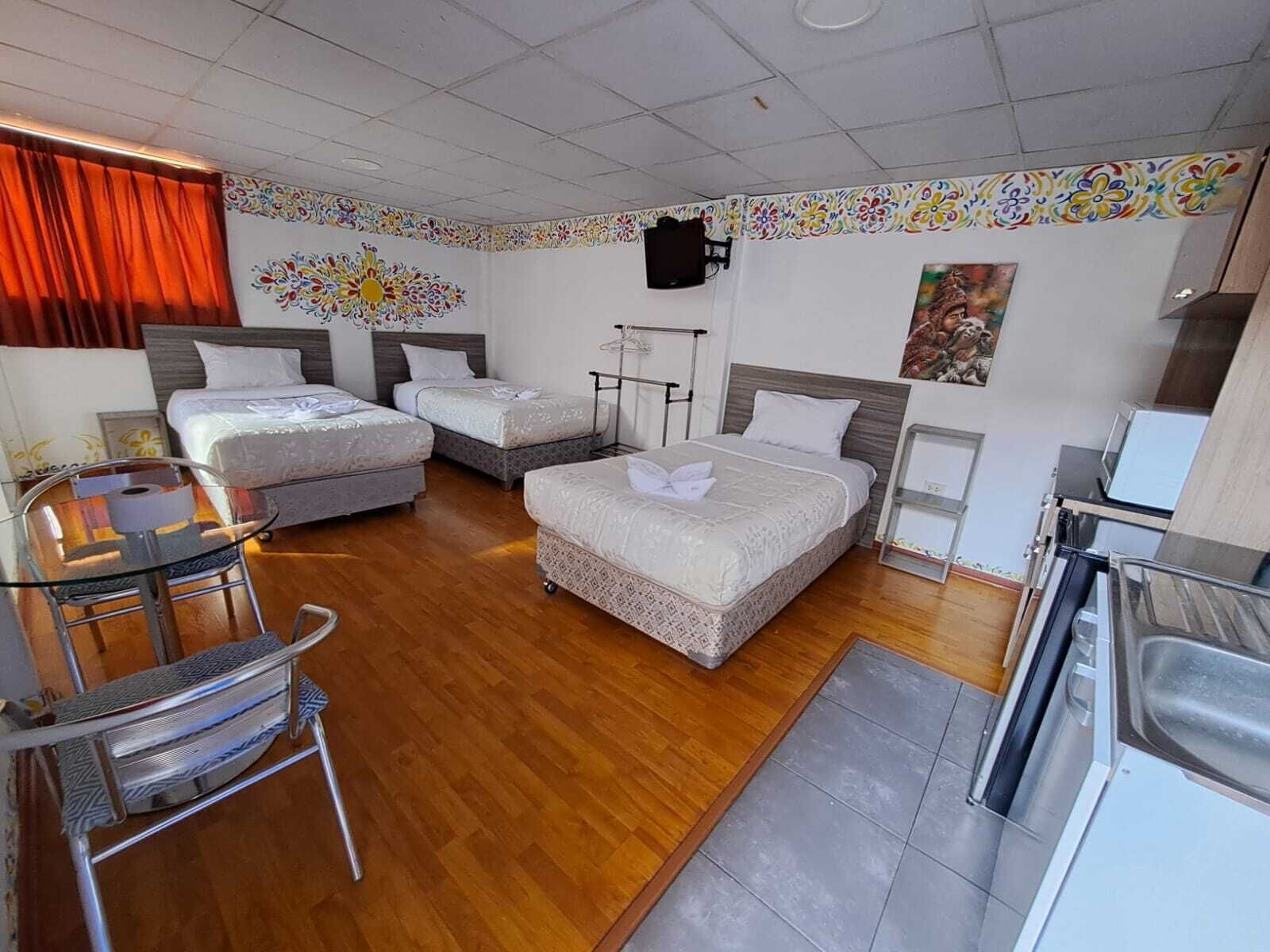 Ett hotellrum med tre sängar, ett litet pentry, ett matbord med stolar och en tv som är monterad på väggen.