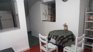 A small room with a white wall, a window, a white chair, a small table with a patterned tablecloth, and a bookshelf. - munay apartment (Cusco)