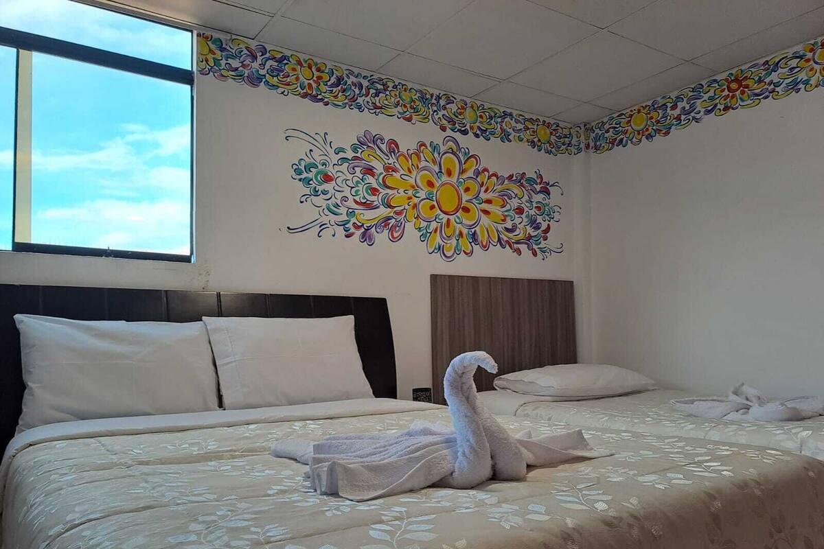 A hotel room with a large bed, a headboard, a window, and a wall with a colorful floral mural.