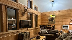 L’intérieur chaleureux d’un chalet, avec un décor en bois, une tête d’animal empaillée accrochée au mur, un téléviseur, une table à manger et une cheminée.