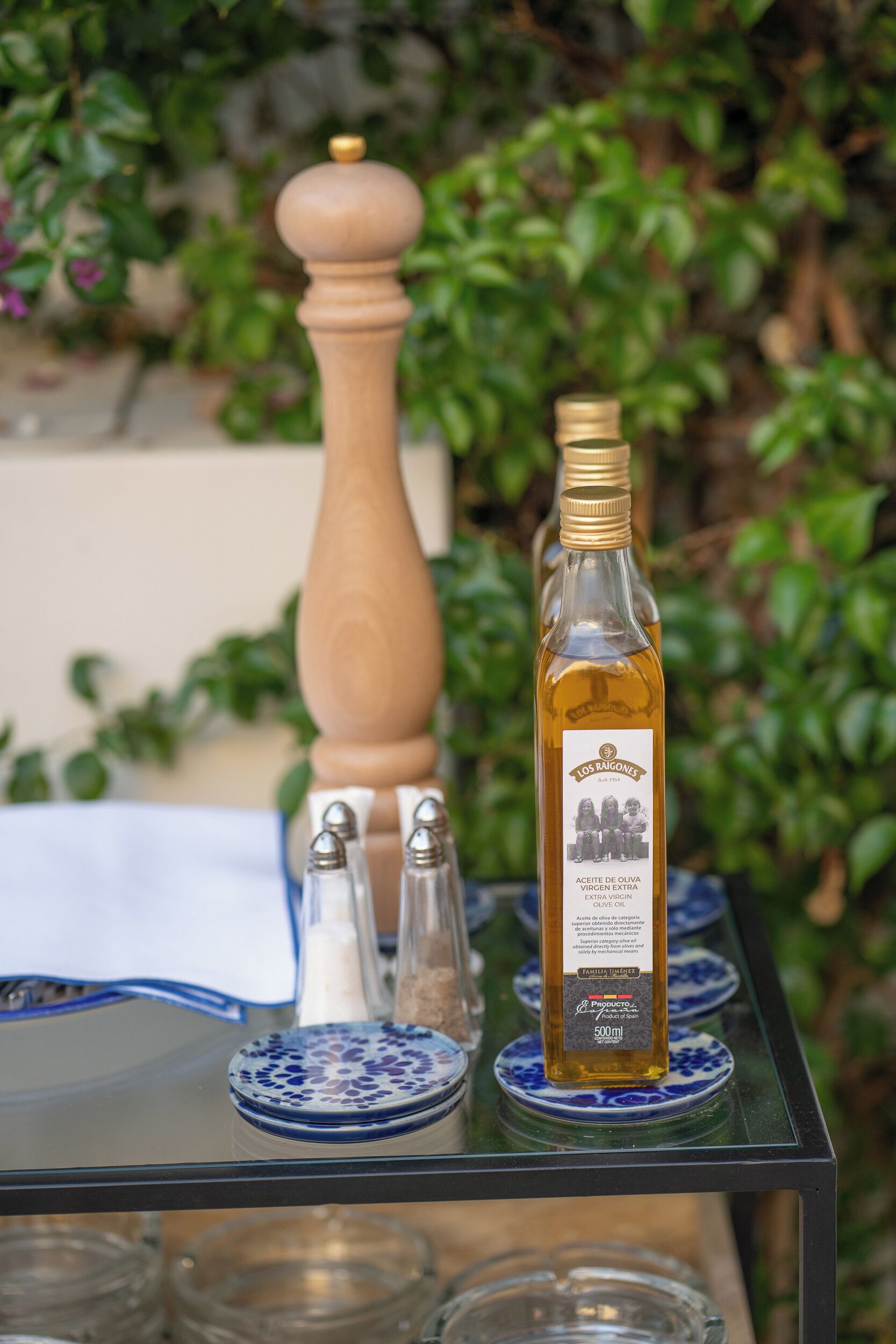 Une bouteille d’huile d’olive est posée sur une table en verre, entourée d’assiettes bleues et blanches à motifs, ainsi que de salières et de poivrières.