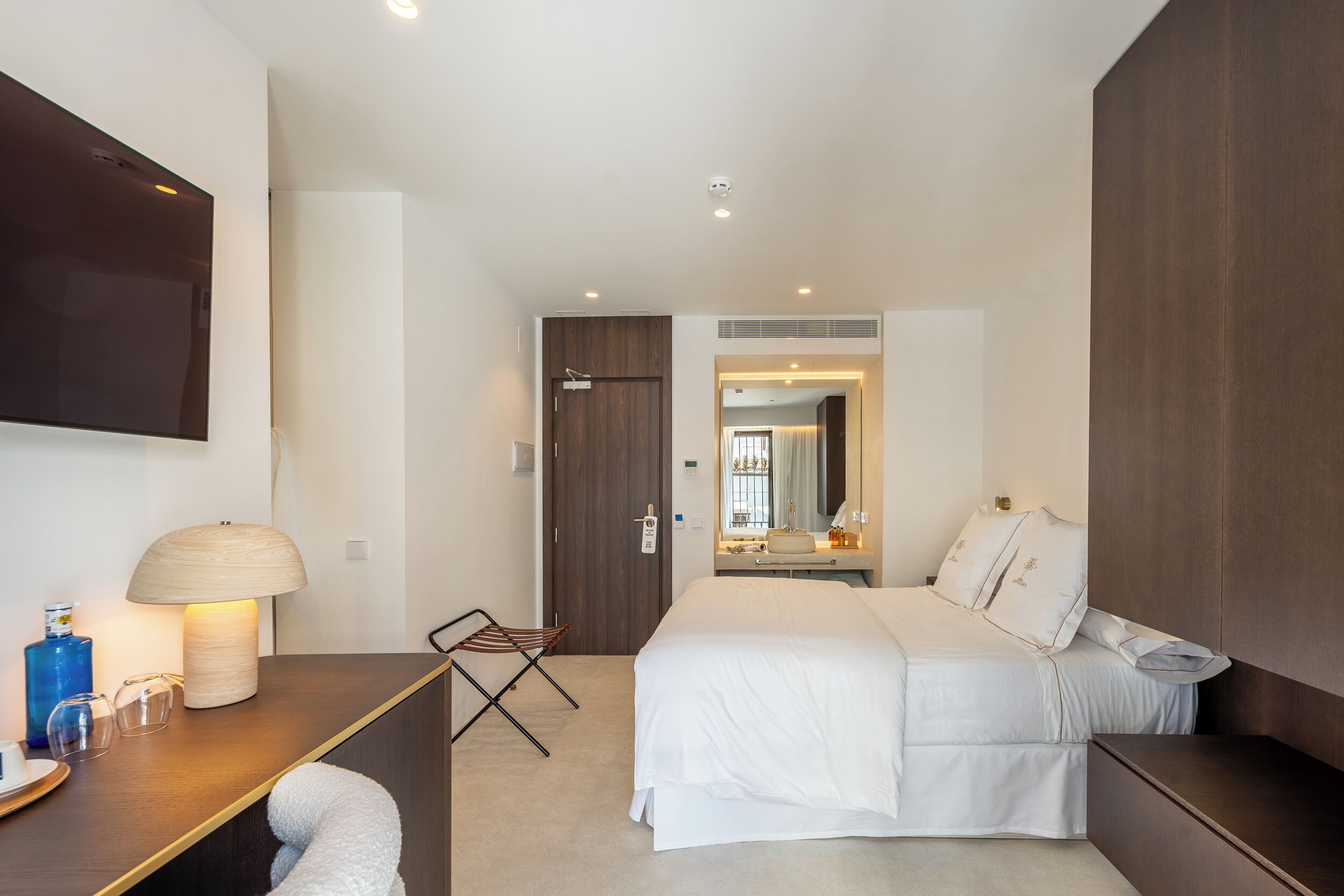 A modern hotel room with a large bed, a desk with a lamp, a flat-screen TV, and a small seating area.