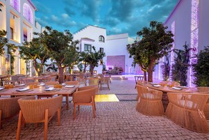 An outdoor dining area with wicker chairs and tables, surrounded by trees and buildings with illuminated windows.