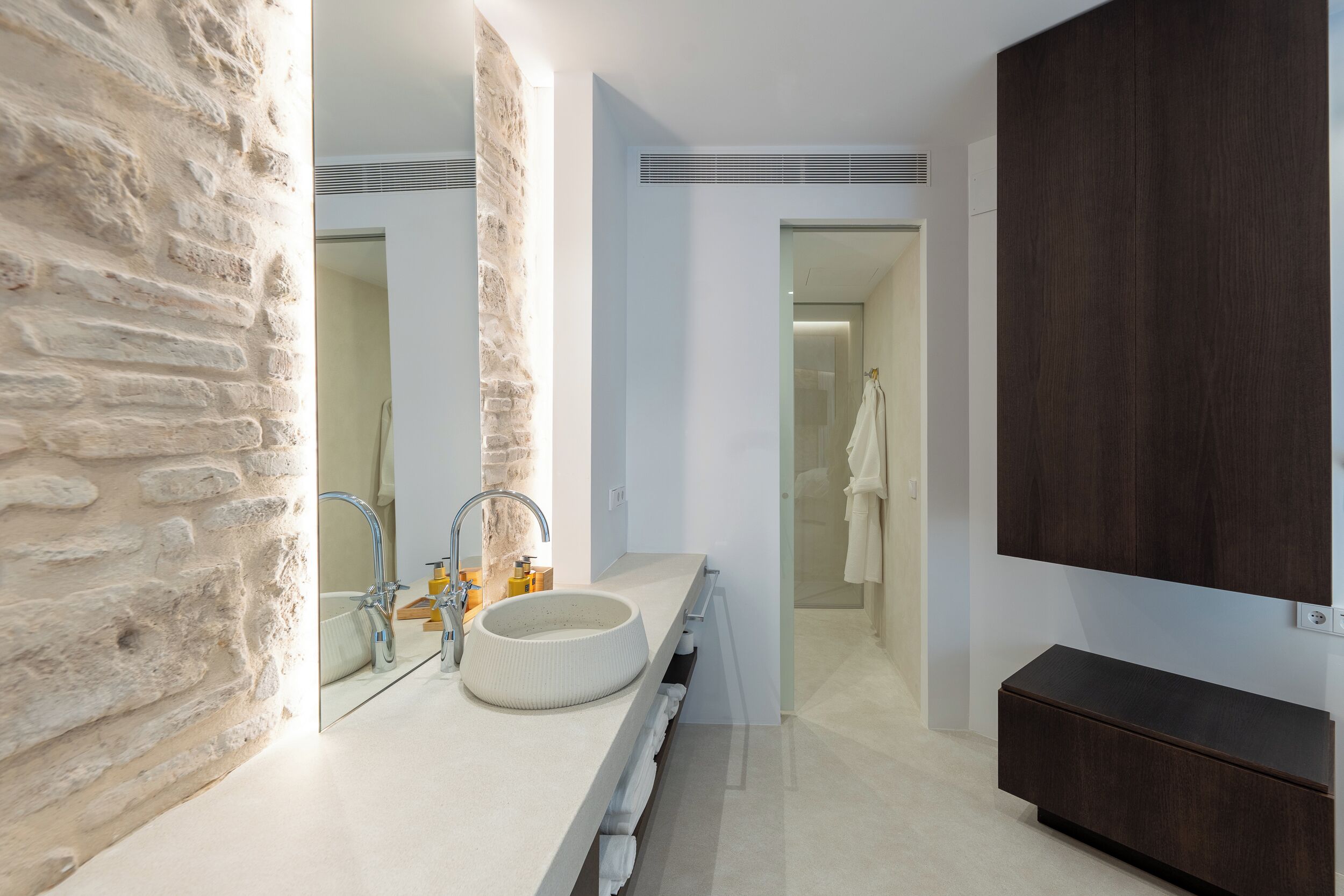 A modern bathroom with a stone wall, a round sink, a mirror, and a dark wood cabinet.