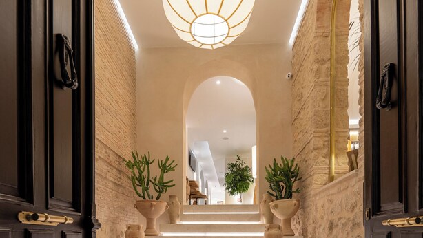 A grand staircase with a modern light fixture, flanked by dark wooden doors and stone walls, leading to a bright interior space.