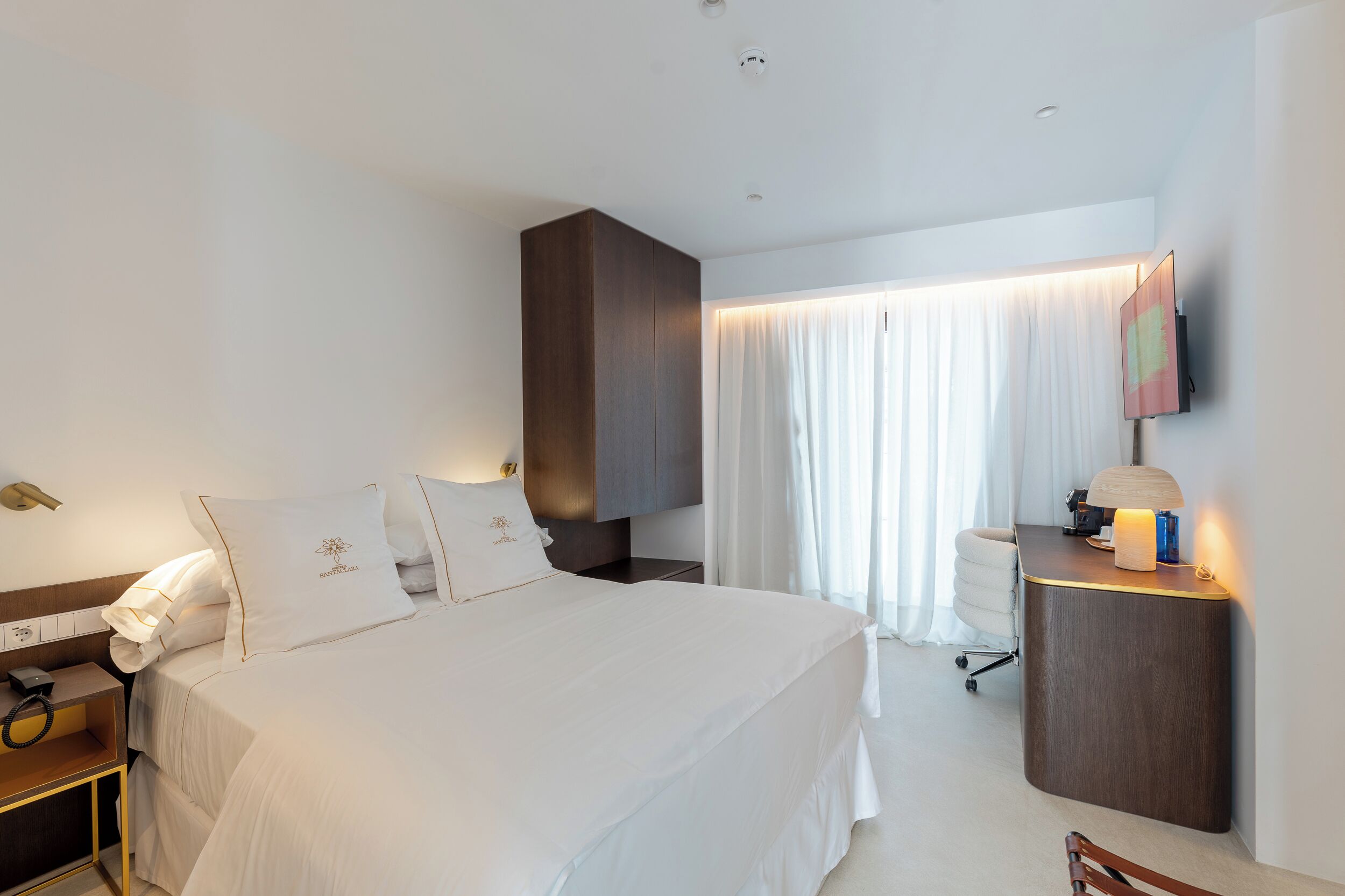 Habitación de hotel con una cama grande, una mesita de noche con lámpara, un escritorio con computadora y una silla.