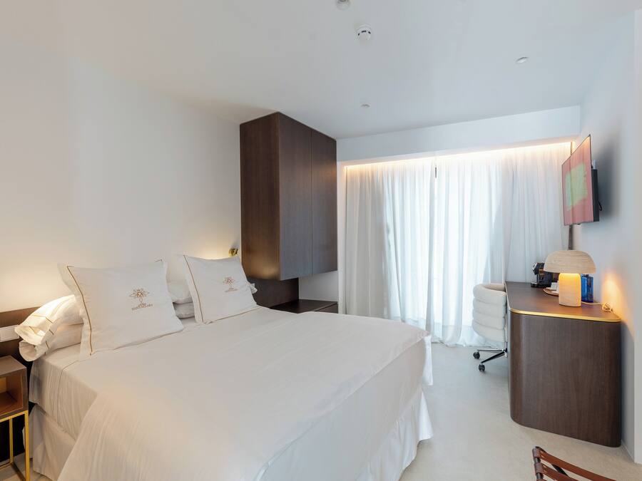 Habitación de hotel con una cama grande, una mesita de noche con lámpara, un escritorio con computadora y una silla.