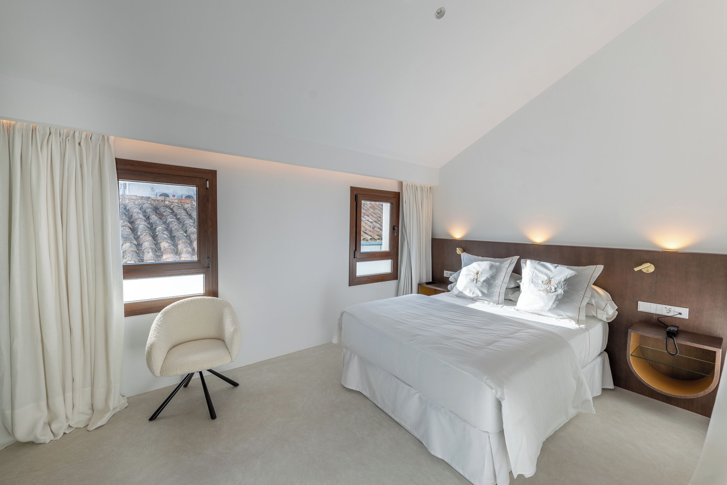 Un dormitorio con cama, una silla, una mesita de noche, un estante en la pared y dos ventanas con cortinas.
