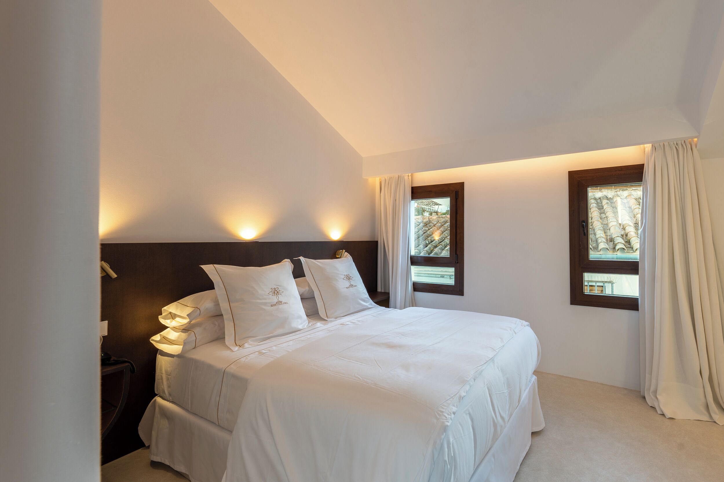 Un dormitorio con cama, mesitas de noche y dos ventanas con cortinas.