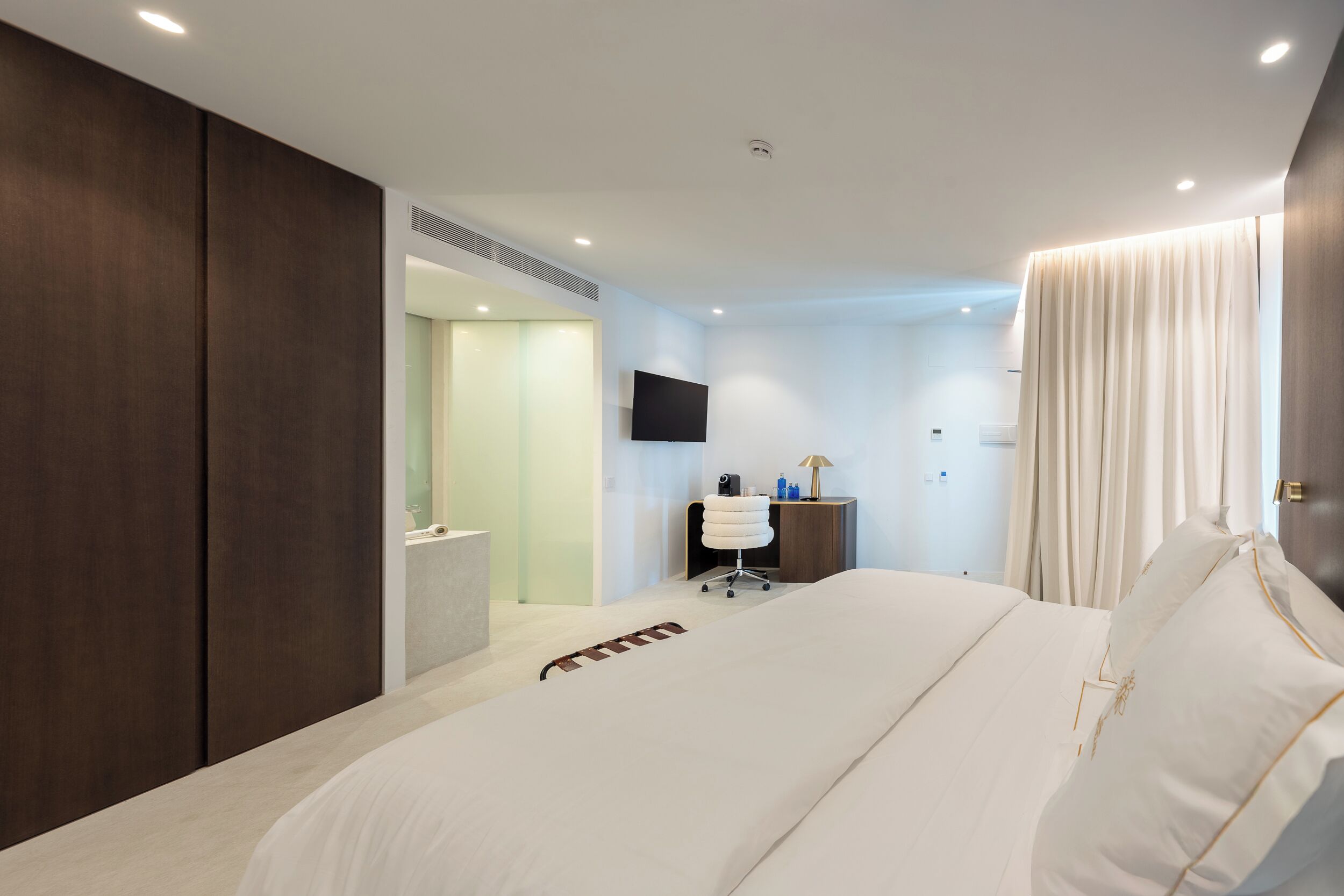 A modern hotel room with a large bed, a desk with a chair, a flat-screen TV, and a bathroom visible through an open door.