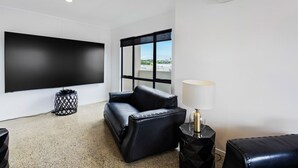 Una sala moderna con un televisor de pantalla plana grande, un sofá de cuero negro, una mesita auxiliar con una lámpara y una ventana con vistas a la ciudad.