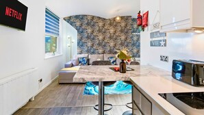 A modern kitchen with a marble countertop, a built-in oven, and a toaster. There is a dining area with a sofa and a small table. A large floral-patterned wall is visible in the background. - Brighton Apartment Near Beach| Walk Everywhere (Brighton and Hove)