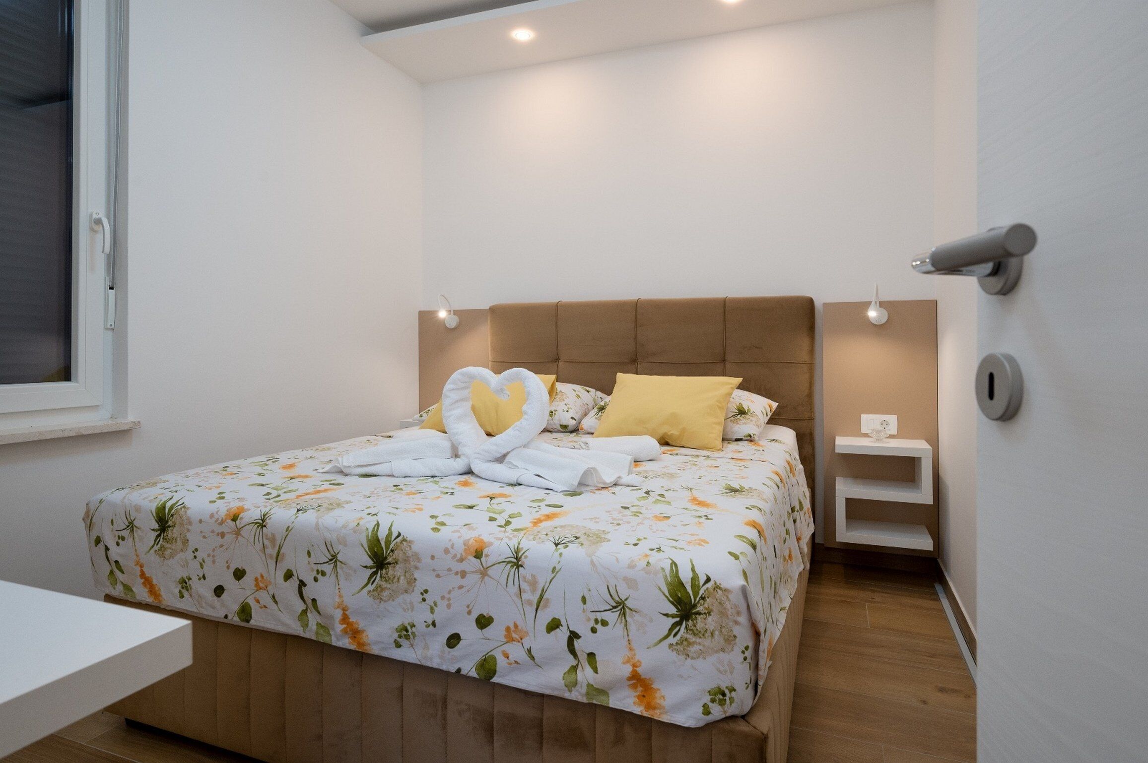 Ein ordentlich bezogenes Bett mit einem blumigen Überwurf und herzförmigen Handtüchern, ein Nachttisch sowie eine an der Wand befestigte Lampe.