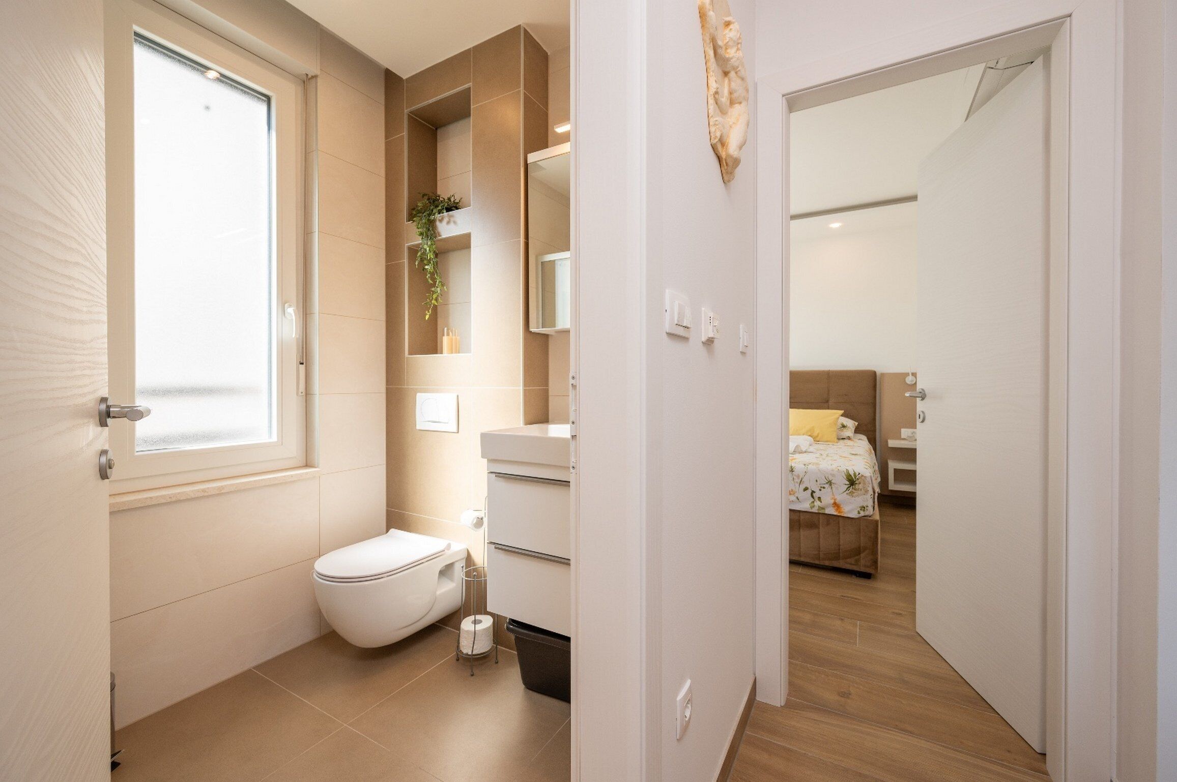 A modern bathroom with a toilet, sink, and mirror. There is a window providing natural light and a view of the bedroom.
