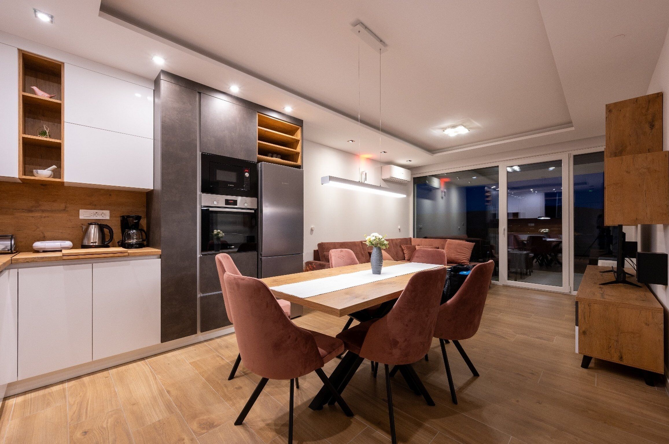 A modern kitchen with a dining area, featuring a wooden table, pink chairs, and a built-in oven.