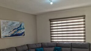 Un salon d’angle avec un canapé d’angle gris, un luminaire au plafond, une fenêtre avec des stores et une œuvre d’art murale qui ressemble à du marbre.