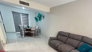 Un salon comprenant un canapé d’angle, un coin repas avec une table et des chaises, et un élément décoratif fixé au mur.