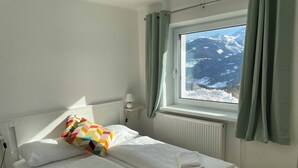 A neatly made bed with white linens and a colorful pillow, a bedside lamp, and a window with a view of snow-covered mountains. - Mountain Hostel (Ramsau am Dachstein)