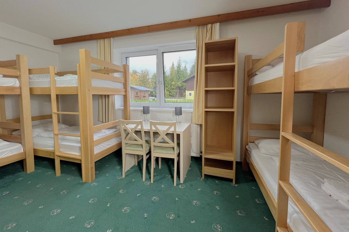 A dormitory room with bunk beds, a table, chairs, and a window with a view of houses.