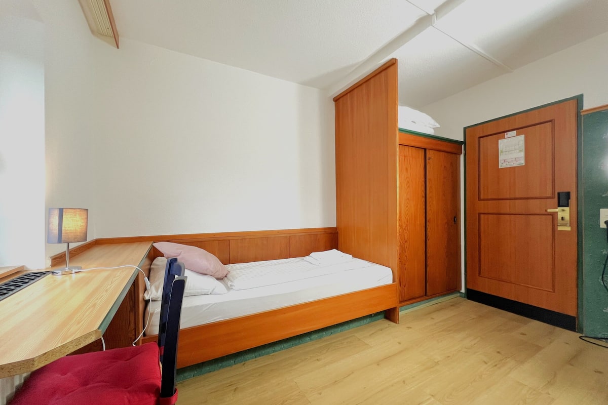 A small, single-bed room with a wooden bed frame, a desk with a lamp, and a wardrobe.