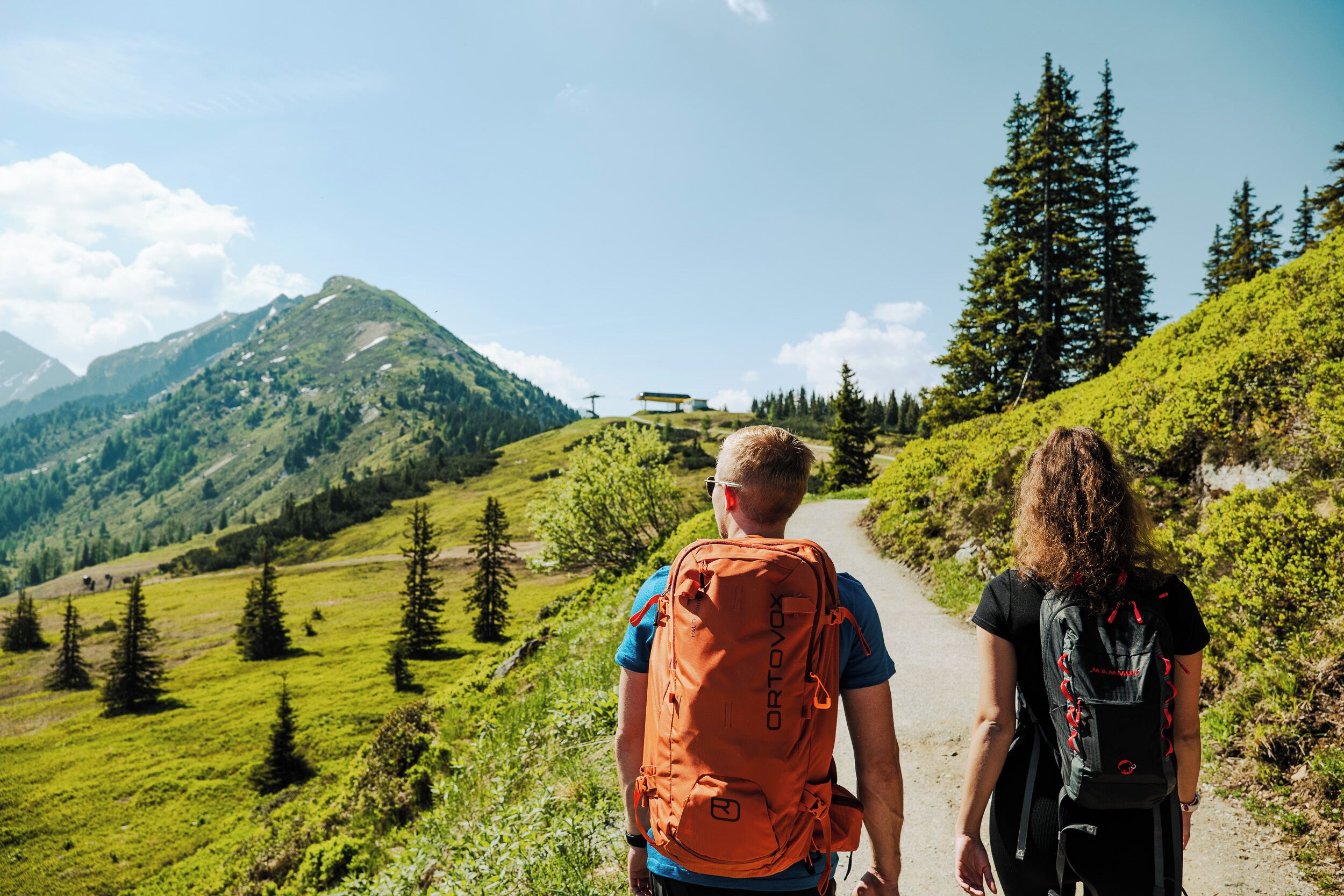 To vandrere på en bjergtrail under en klar himmel og frodigt grønt landskab.
