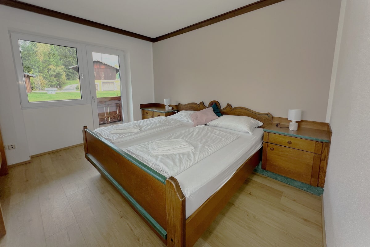 A bedroom with a wooden bed, a nightstand, and a window with a view of greenery.