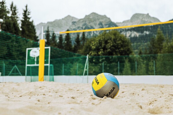Una pelota de voleibol sobre una cancha de arena con una red, rodeada por una cerca verde y montañas al fondo.