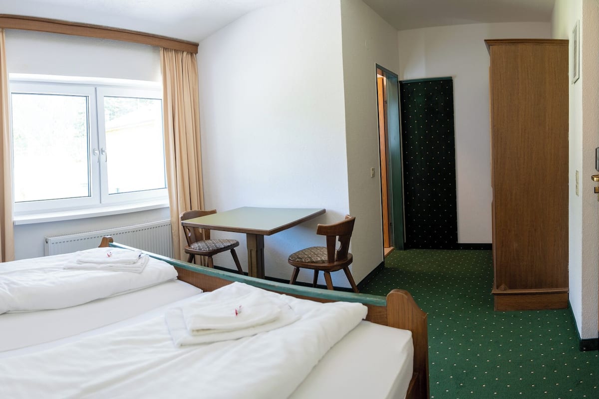 A hotel room with two beds, a small table with chairs, a wardrobe, and a view of the outdoors through a window.