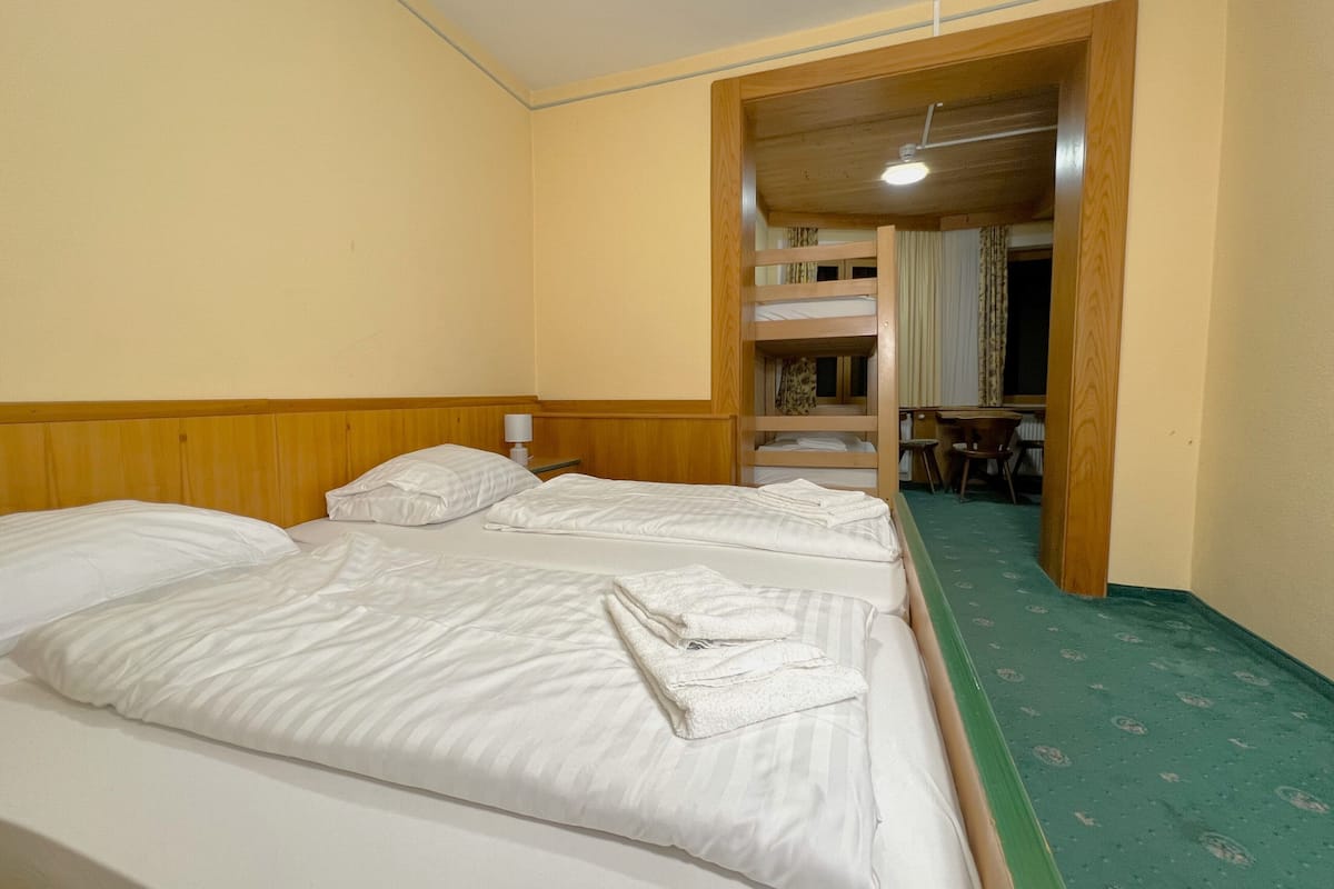 A hotel room with two beds, a wooden headboard, a desk, and a bunk bed in the background.