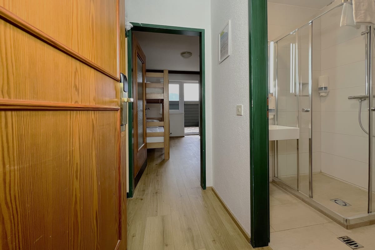 A hotel corridor with a wooden door, a glass shower area, and a view into a room with a bunk bed.
