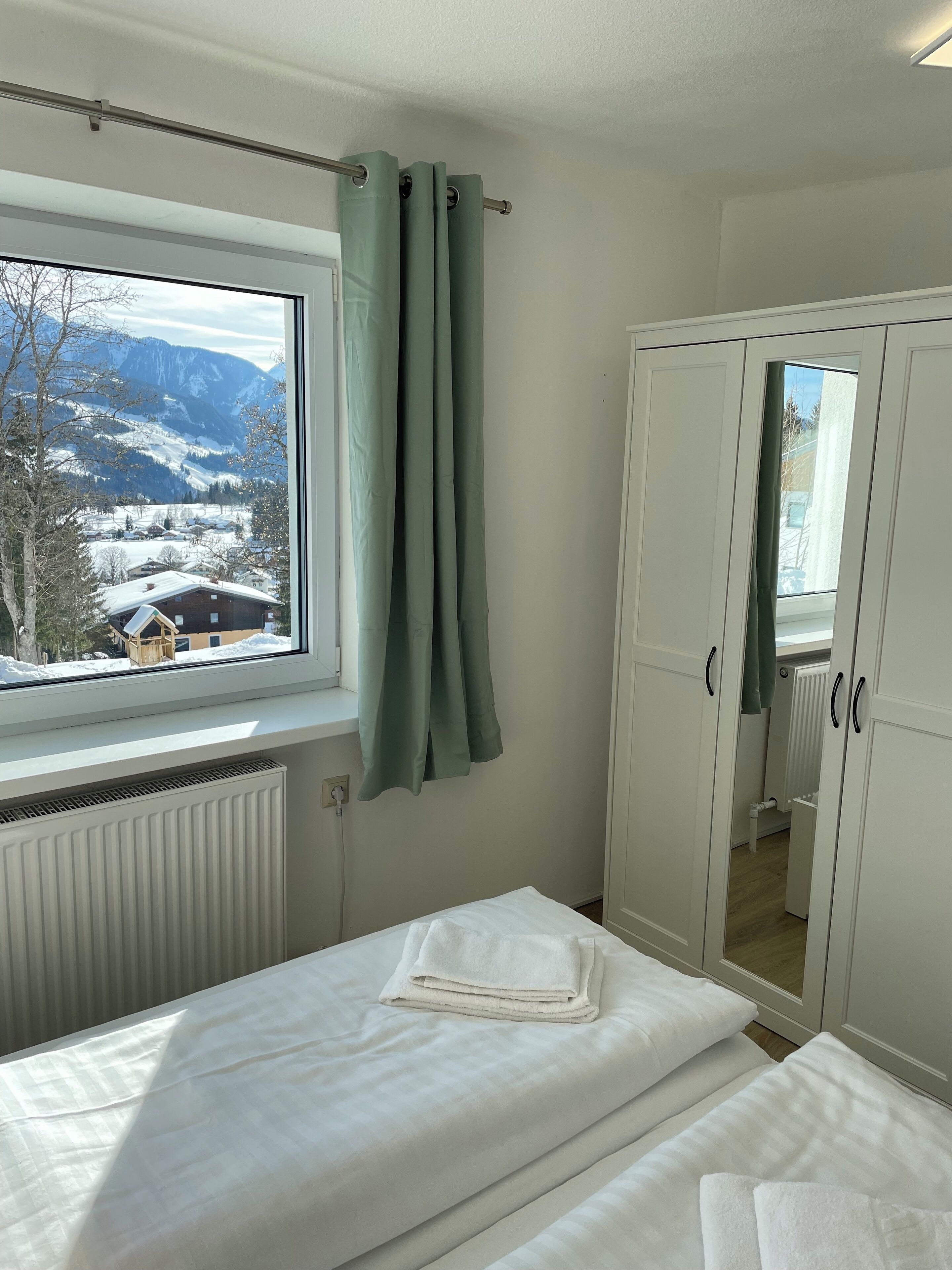 A bedroom with a bed, a window with curtains, a wardrobe, and a view of snow-covered mountains.