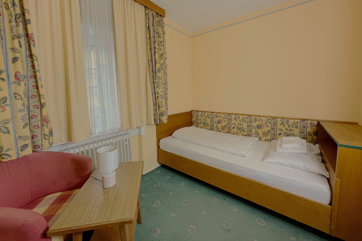 A small, single-room hotel room with a bed, a chair, a small table, and a window with curtains.