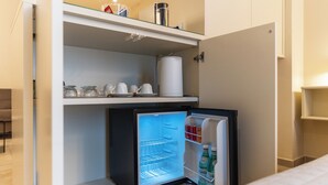 A compact refrigerator with a freezer compartment, a shelf above containing glass jars and a kettle, and a door shelf with bottled beverages. - Cozy studio apartment in the center of Bari, renovated and tastefully furnished (Bari)