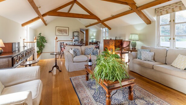 A spacious living room with a high ceiling and exposed wooden beams, a large window with curtains, a sofa set, a coffee table with a plant, and a dining area in the background. - Buckstone House Coach House, Forest of Dean - sleeps 4 guests in 2 bedrooms (Forest of Dean)
