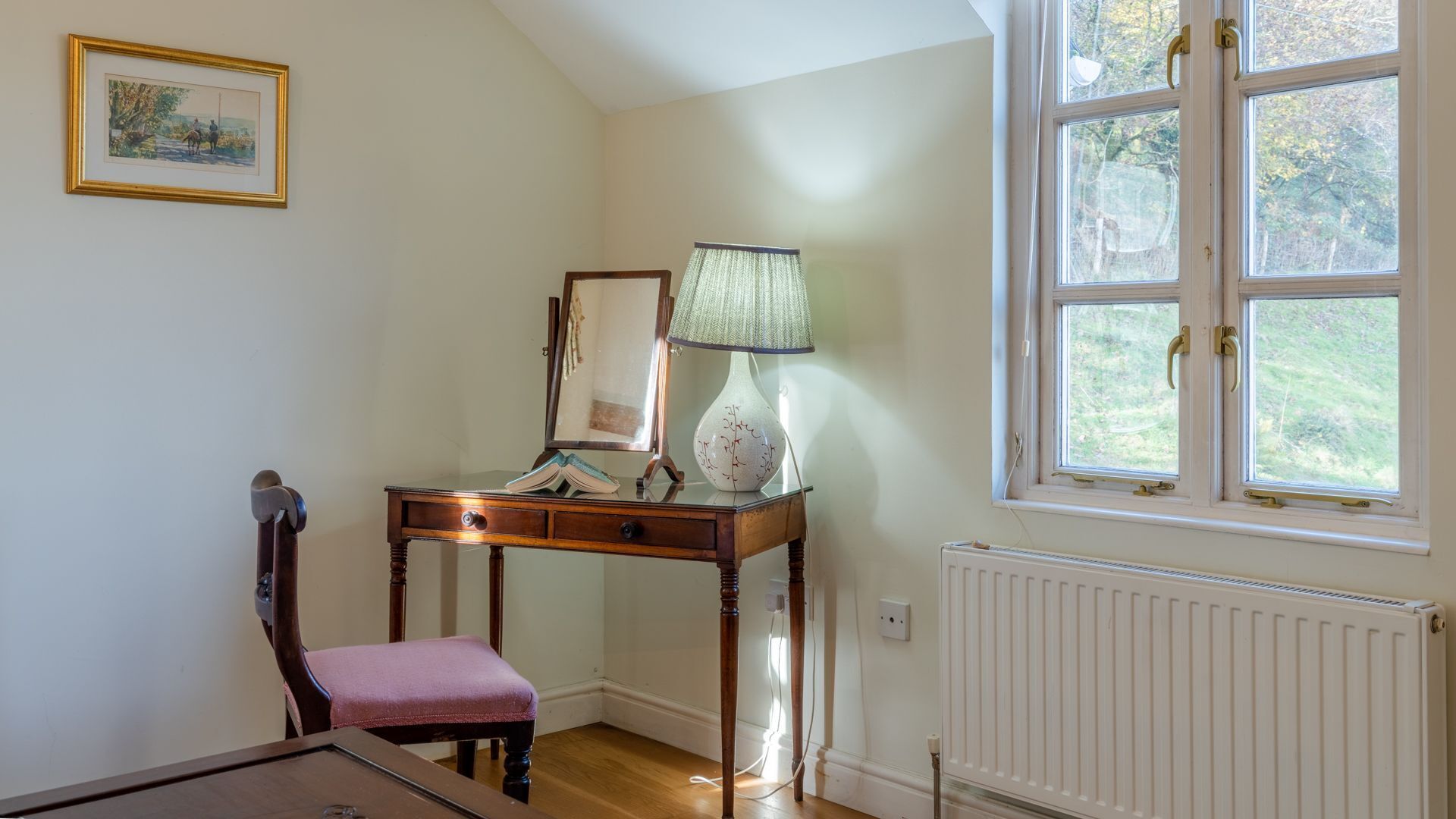 A room with a wooden desk, a chair, a lamp, a mirror, and a window with a view of greenery.