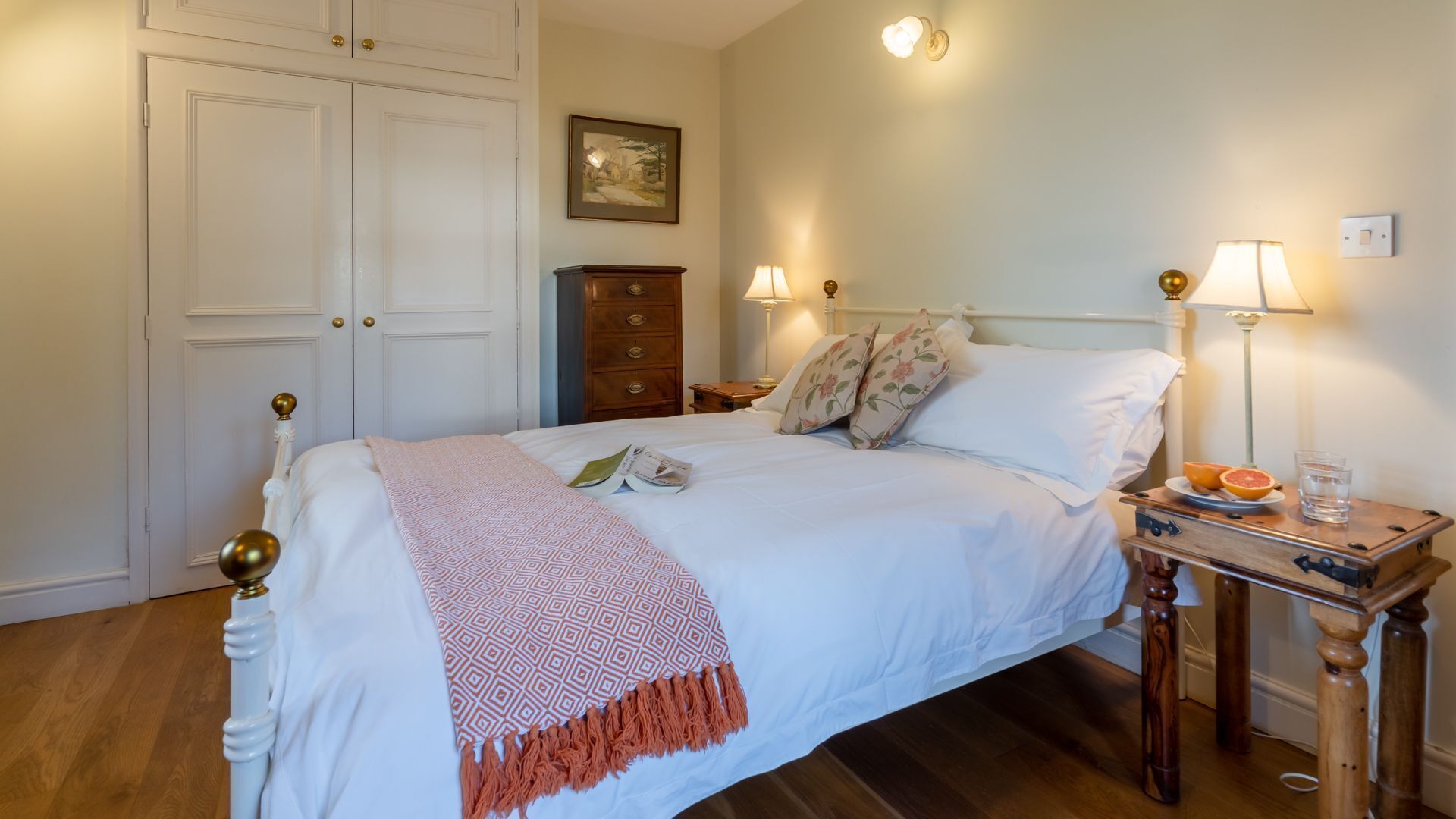 Un lit bien fait, recouvert d’un couvre-lit à motifs, une table de chevet avec une lampe et des fruits, et une commode avec des tiroirs.