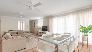 A modern living room with a glass dining table, white furniture, a ceiling fan, and a large window with sheer curtains. - Noosa International 145 Noosa Heads (Noosa Heads)