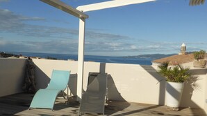 Una terraza con dos sillones de descanso, una planta en maceta y vistas al mar y edificios a la distancia.
