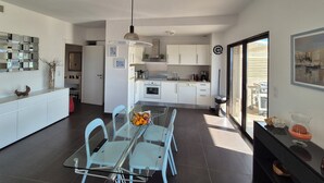 Una cuina moderna amb armaris blancs, una zona de menjador amb taula de vidre, i una finestra gran.