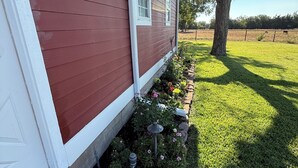 A red house with a white door and windows, a garden with flowers and a tree, and a clear sky. - Cozy 1-bedroom apartment in charming Burton 9 minutes from ROUND TOP! (Burton)