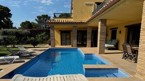 Une piscine avec des chaises longues, un espace patio avec une table et des chaises, et un bâtiment à la façade jaune et aux piliers en pierre.