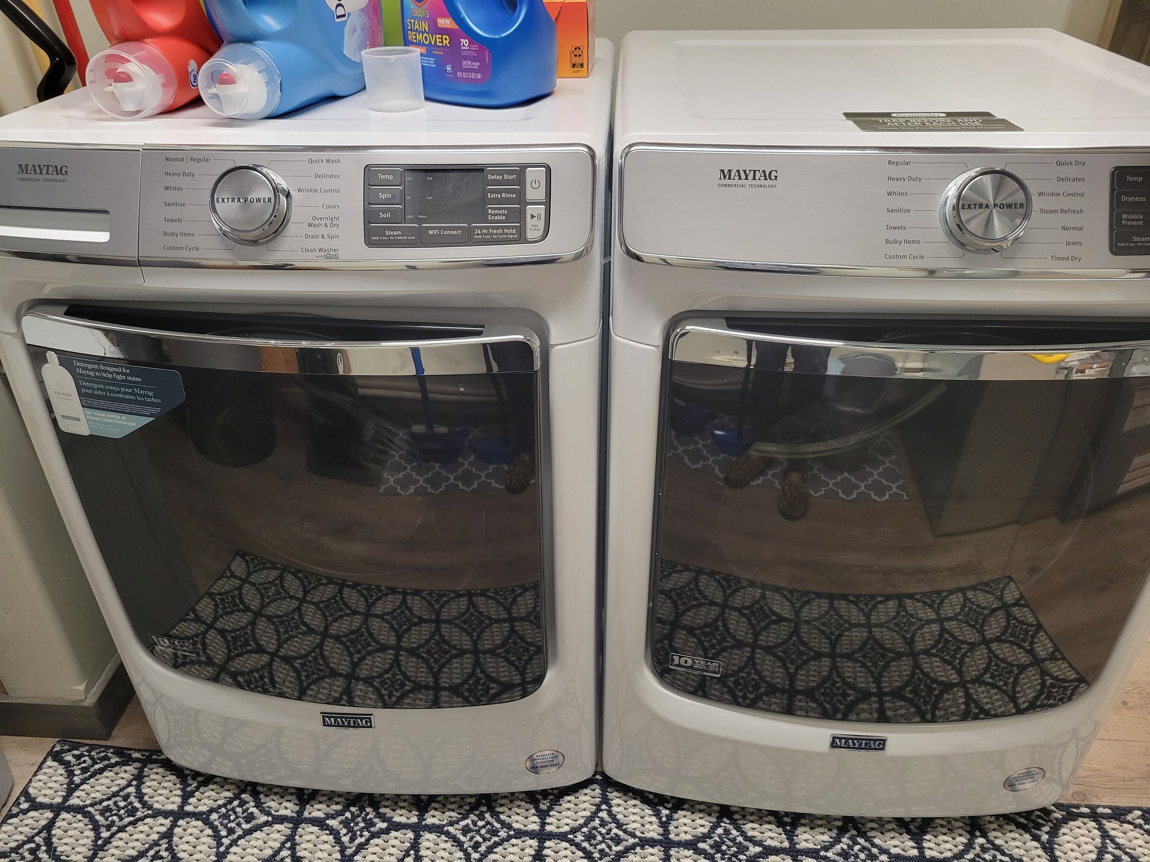 Two Maytag front-loading washing machines with control panels, a small table with cleaning supplies, and a patterned rug on the floor.