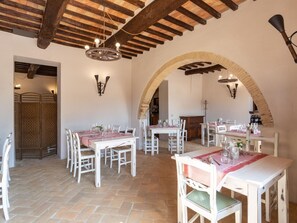 Un comedor con vigas de madera rústicas, mesas y sillas blancas. Hay dos candelabros y apliques de pared. Un arco de entrada conduce a otra habitación.