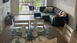 A dining area with a wooden table set for four, a black sofa with a throw blanket, and a large window with a view of a garden and a building. - Beautiful Resort Near Theme Parks (Kissimmee)