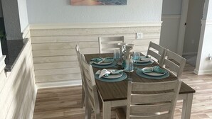 A dining area with a wooden table set for four, surrounded by light-colored chairs. A painting of a sunset over the ocean hangs on the wall above the table. - Beautiful Resort Near Theme Parks (Kissimmee)